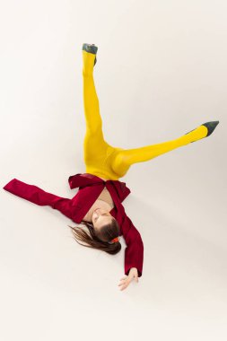 Sarı taytlı, kırmızı ceketli, bacakları havada yerde yatan, gri stüdyo arka planında yalnız poz veren şık bir kız. Tuhaf bir ruh hali. Eski moda kavramı, sanat fotoğrafçılığı, stil, nonoş, güzellik.