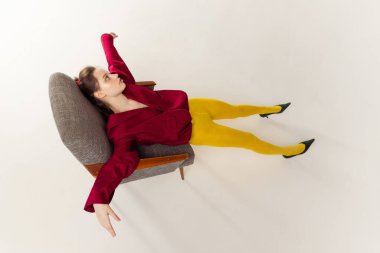 Portrait of young girl in yellow tights and red jacket posing, lyying on chair isolated over grey studio background. Concept of retro fashion, art photography, style, queer, beauty