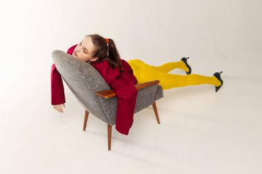 Portrait of young girl in yellow tights and red jacket posing with chair isolated over grey studio background. Extraordinary pose. Concept of retro fashion, art photography, style, queer, beauty