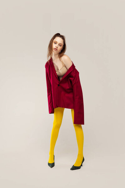 Portrait of young stylish girl in yellow tights and red jacket posing isolated over grey studio background. Extravagant bright outfit. Concept of retro fashion, art photography, style, queer, beauty