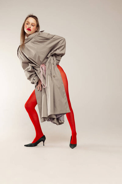 Portrait of young stylish girl posing in red tights and coat posing isolated over grey studio background. Red lipstick look. Concept of retro fashion, art photography, style, queer, beauty