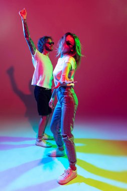 Portrait of young stylish man and woman dancing, posing isolated over red background in neon light. Dancing. Feeling good. Concept of youth, lifestyle, relationship, emotions, facial expression, ad