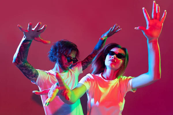 Portrait of young stylish man and woman dancing, posing isolated over red background in neon light. Emotive party time. Concept of youth, lifestyle, relationship, emotions, facial expression, ad