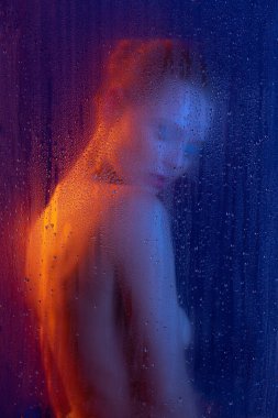 Parlak makyajlı genç bir kadının portresi mavi arka planda, neon ışıkta izole edilmiş şekilde duruyor. Sisli cam etkisi. Sisli cam etkisi. Güzellik, moda, tarz, kozmetoloji, kozmetik kavramı.