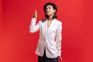 Idea. Portrait of stylish young woman in black beret and white jacket posing isolated over red studio background. Concept of beauty, fashion, style, emotions, femininity, facial expression, sales, ad