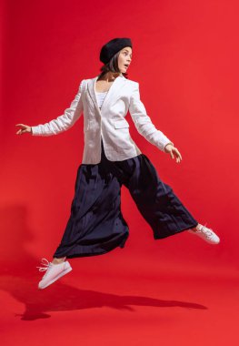 Kırmızı stüdyo arka planında izole edilmiş siyah bereli ve beyaz ceketli şık bir genç kadının portresi. Atlıyorum. Güzellik, moda, tarz, duygu, kadınlık, yüz ifadesi, satış, reklam