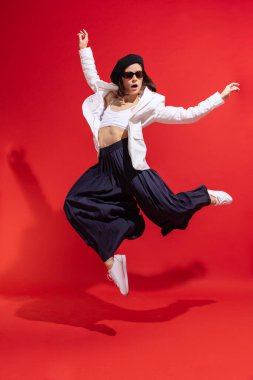 Stylish young woman in black beret and sunglasses posing, dancing isolated over red studio background. Concept of beauty, fashion, style, emotions, femininity, facial expression, sales, ad