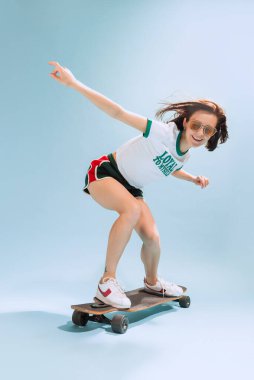 Portrait of happy woman in casual summer outfit posing on skateboard isolated over light blue background. Active hobby. Concept of beauty, style, emotions, fashion, lifestyle, facial expression, ad