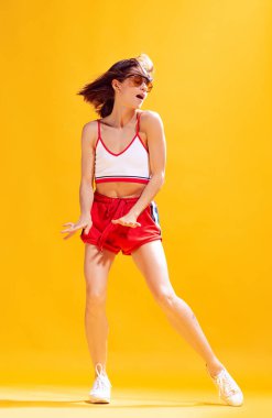 Üstteki güzel genç kadın portresi ve sarı arka planda izole edilmiş kırmızı şort. Neşeli bir dans. Güzellik kavramı, tarz, duygu, moda, yaşam tarzı, yüz ifadesi, reklam