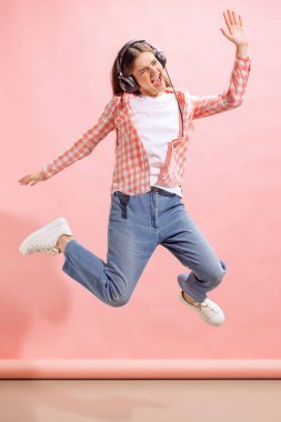 Portrait of emotive young girl in checkered shirt listening to music in headphones and jumping isolated over pink studio background. Concept of youth, fashion, lifestyle, emotions, facial expression.