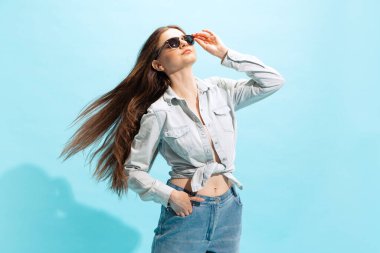 Mavi stüdyo arka planında izole edilmiş günlük kıyafetler ve güneş gözlüğü takmış güzel bir genç kızın portresi. Gençlik kavramı, moda, yaşam tarzı, duygular, yüz ifadesi. İlan için alanı kopyala