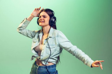 Portrait of young emotive girl in casual clothes listening to music in headphones isolated on green studio background. Catching vibe. Concept of youth, fashion, lifestyle, emotions, facial expression.