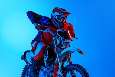 Yoğunlaşmış genç bir adamın portresi, çapraz motosiklet kullanan motosikletli bir adam mavi stüdyo arka planında, neon ışıklarıyla izole edilmiş. Motosport, hız, aksiyon, hobi, rekabet kavramı. İlan için alanı kopyala