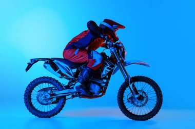 Genç bir adamın portresi, motosikletli tam teçhizatlı bir motosikletçi mavi stüdyo arka planında, neon ışıkta izole edilmiş. Motokros. Motosport, hız, aksiyon, hobi, rekabet kavramı. İlan için alanı kopyala