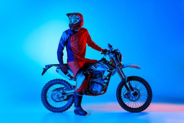 Genç bir adamın portresi, profesyonel spor ekipmanları içinde motosikletçi mavi stüdyo arka planında, neon ışıkta izole edilmiş. Dinleniyor. Motosport, hız, aksiyon, hobi, rekabet kavramı. İlan için alanı kopyala