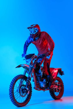 Genç bir adamın portresi, ekipmanlı motosikletçi mavi stüdyo arka planında, neon ışıkta izole edilmiş. Motokros yarışması. Motosport, hız, aksiyon, hobi, rekabet kavramı. İlan için alanı kopyala