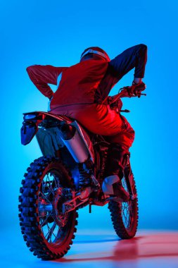 Motosiklet üzerinde poz veren genç bir adamın portresi mavi stüdyo arka planında neon ışıkta izole edilmiş. Arkadan bak. Motosport, hız, aksiyon, hobi, rekabet kavramı. İlan için alanı kopyala