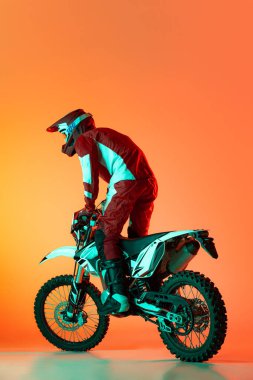 Genç bir adamın portresi, motosiklet kullanan tam teçhizatlı bir motosikletçi turuncu stüdyo arka planında neon ışıklarıyla izole edilmiş. Dönüyor. Motosport, hız, aksiyon, hobi, rekabet kavramı. İlan için alanı kopyala