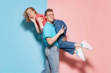 Neşeli genç bir çiftin portresi, omuzlarında kadını taşıyan bir adam pembe mavi stüdyo arka planında izole edilmiş. Gençlik kavramı, duygular, yüz ifadesi, aşk, ilişki. Poster, reklam