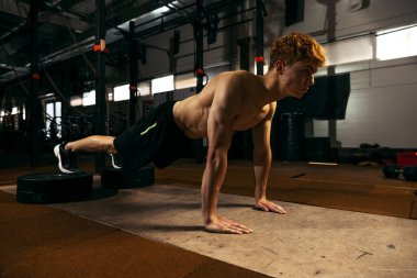 Kaslı vücut eğitimi almış genç kızıl saçlı sporcunun portresi geym arka planında izole edilmiş şınav egzersizleri yapıyor. Spor, sağlık, eylem, beslenme, gençlik kavramı. İlan için alanı kopyala