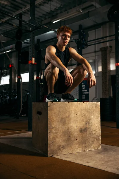 Portrait of sportive, muscular red-haired young man sitting on box, resting isolated over gym background. Sweaty. Concept of sport, health, action, nutrition, youth. Copy space for ad