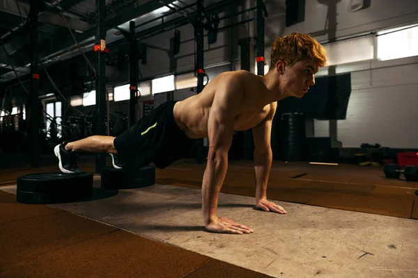 Portrait of young red-haired sportive man with muscular body training, doing push-ups exercises isolated over geym background. Concept of sport, health, action, nutrition, youth. Copy space for ad