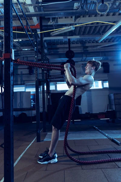 Full-length portrait of muscular red-haired man training, doing exercises with ropes isolated over gym background. Concept of sport, health, action, nutrition, youth. Copy space for ad