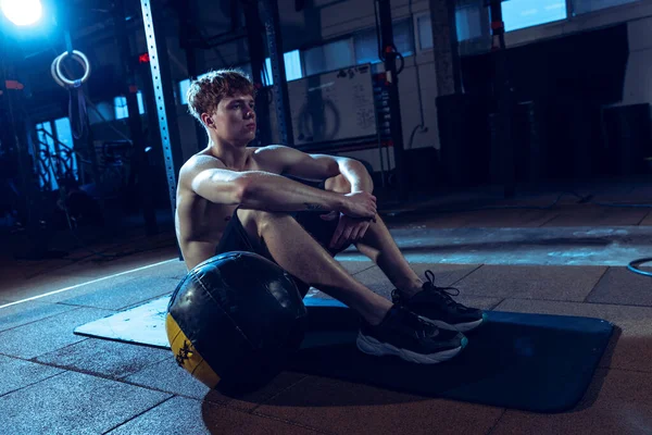 Portrait of young sportive man sitting on fitness rug isolated over gym background. Resting. Workout routine. Concept of sport, health, action, nutrition, youth. Copy space for ad