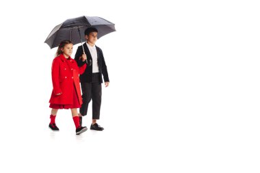 İki çocuğun portresi, siyah takım elbiseli bir oğlan ve kırmızı ceketli bir kız beyaz arka planda şemsiye altında yürüyor. Çocukluk kavramı, arkadaşlık, eğlence, yaşam tarzı, moda. Retro biçimi