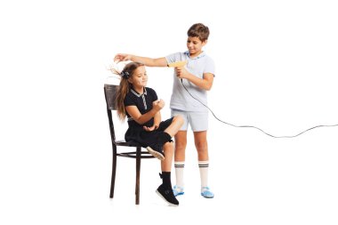 Çocukların portresi, beyaz stüdyo arka planında izole edilmiş saç kurutma makinesiyle kızların saçını kurutmaya yardım eden çocuk. Çocukluk kavramı, arkadaşlık, eğlence, yaşam tarzı, moda. Retro biçimi