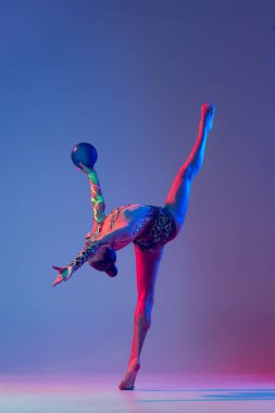 Portrait of young girl, rhythmic gymnast athlete training with blue ball isolated over purple studio background in neon light. Twine. Concept of action, motion, sport life, motivation, competition.