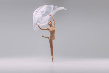 Portrait of young beautiful ballerina dancing with transparent fabric isolated over grey studio background. Grace. Concept of classic ballet, inspiration, beauty, dance, creativity