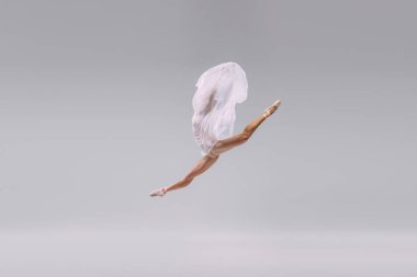 Portrait of young ballerina dancing, jumping in a twine with transparent fabric isolated over grey studio background. Concept of classic ballet, inspiration, beauty, dance, creativity