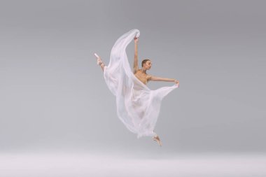 Portrait of young beautiful ballerina dancing, jumping with transparent fabric isolated over grey studio background. Concept of classic ballet, inspiration, beauty, dance, creativity
