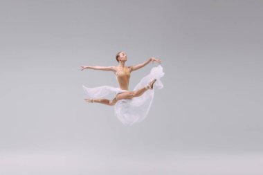 Portrait of young ballerina dancing with light transparent fabric isolated over grey studio background. Flying like a bird. Freedom. Concept of classic ballet, inspiration, beauty, dance, creativity