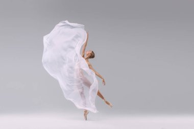 Portrait of young ballerina dancing with fabric isolated on grey studio background. Standing on tiptoe. Weightless transparent cloth. Concept of classic ballet, inspiration, beauty, dance, creativity
