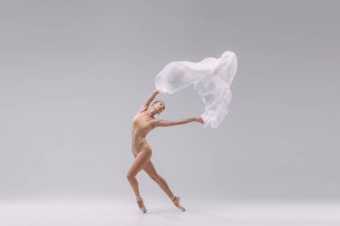 Portrait of young ballerina dancing in beige bodysuit with transparent fabric isolated over grey studio background. Tiptoe movements. Concept of classic ballet, inspiration, beauty, dance, creativity
