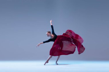 One young ballerina dancing with deep red fabric isolated over blue grey studio background. Artistic performance of classic dance. Concept of classic ballet, inspiration, beauty, dance, creativity