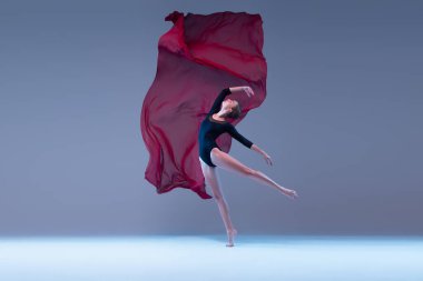 One young tender ballerina dancing with deep red transparent fabric isolated over blue grey studio background. Attractive performance. Concept of classic ballet, inspiration, beauty, dance, creativity