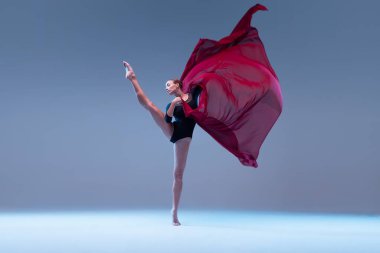 Portrait of young ballerina dancing with deep red fabric isolated over blue grey studio background. Tender classical movements. Concept of classic ballet, inspiration, beauty, dance, creativity