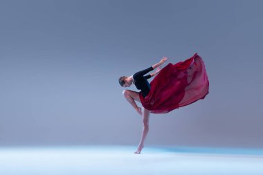 Portrait of young ballerina dancing in black bodysuit with deep red fabric isolated on blue grey studio background. Grace, tenderness. Concept of classic ballet, inspiration, beauty, dance, creativity