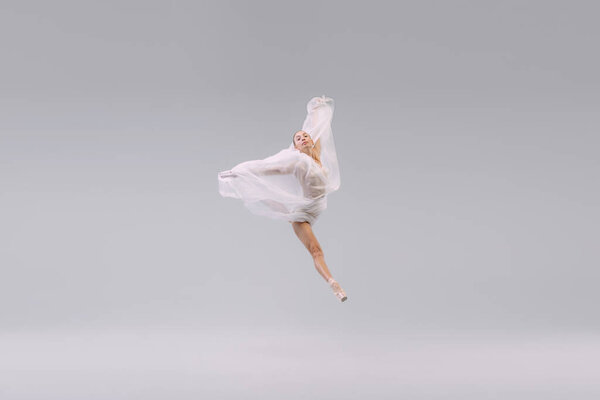 Portrait of young ballerina dancing, jumping with transparent fabric isolated over grey studio background. Leap in the air. Concept of classic ballet, inspiration, beauty, dance, creativity