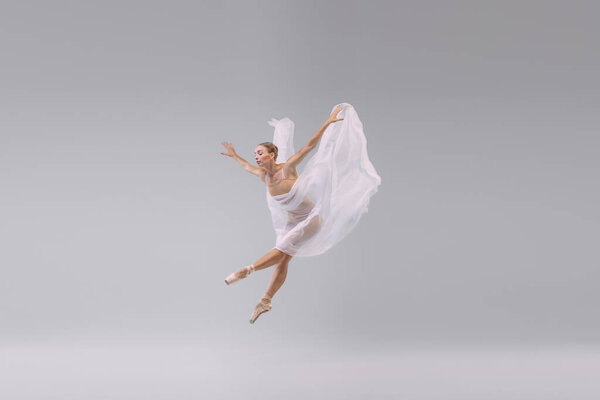 Portrait of young ballerina dancing with fabric isolated over grey studio background. The grace, artist, movement. Fly. Concept of classic ballet, inspiration, beauty, dance, creativity
