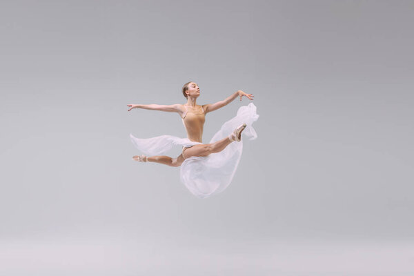 Portrait of young ballerina dancing with light transparent fabric isolated over grey studio background. Flying like a bird. Freedom. Concept of classic ballet, inspiration, beauty, dance, creativity