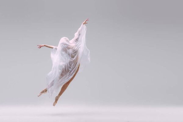 Portrait of young ballerina dancing fully covered with fabric isolated over grey studio background. Flight of dreams. Concept of classic ballet, inspiration, beauty, dance, creativity