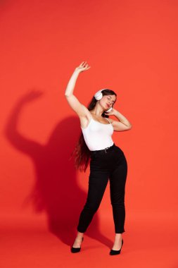 Portrait of beautiful young woman posing, listening to music in headphones, dancing isolated over red studio background. Concept of beauty, lifestyle, youth, emotions, facial expression, ad