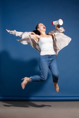 Portrait of young woman posing, shouting in megaphone in a jump isolated over blue studio background. Big sales, Black Friday. Concept of shopping, lifestyle, youth, emotions, facial expression, ad