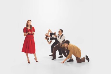 Group of young men in stylish suits taking photo of beautiful woman in red dress isolated over white studio background. Popularity. Concept of retro fashion, style, youth culture, emotions, beauty, ad