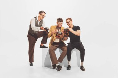 Portrait of three stylish young men in trendy suits sitting, looking at retro camera photos isolated over white studio background. Concept of retro fashion, style, youth culture, emotions, art, ad