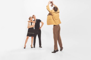 Portrait of man in yellow retro jacket taking photo with vintage camera of beautiful couple isolated over white studio background. Concept of retro fashion, style, youth culture, emotions, beauty, ad